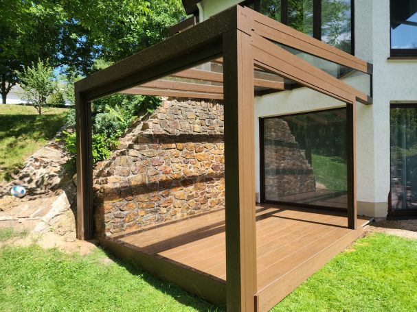 Terrassenüberdachung Moderne Terrasse mit Holzrahmen und Wänden aus Glas, umgeben von Rasen und Bäumen.